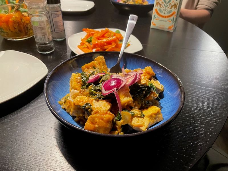 A bowl of Noor Murad's Black Lime Tofu with a sideplate of Roasted Carrot Salad with Chamoy in the background