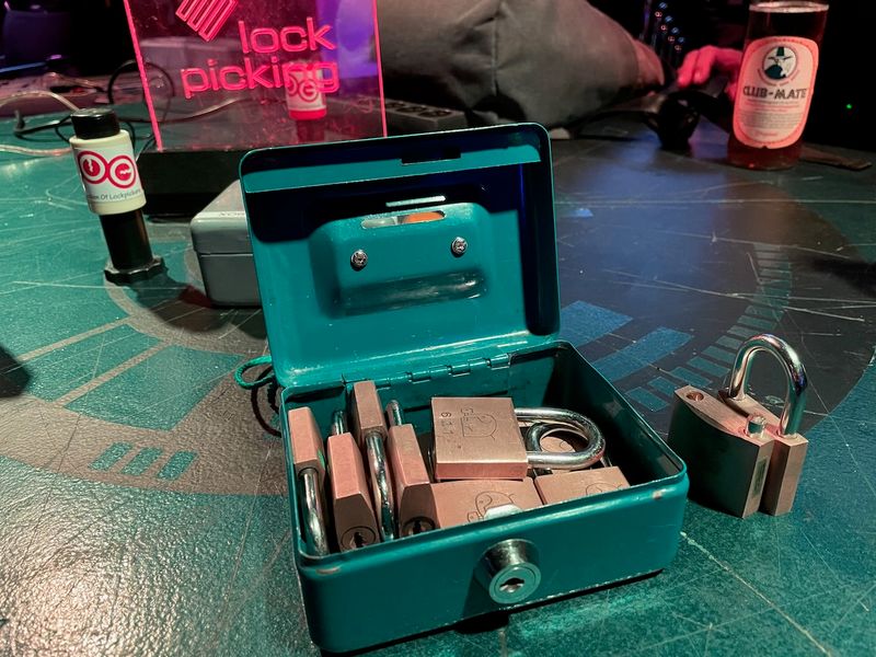 Ten or so locks in a box and on a table. One of the locks is broken. There's also a bottle of Club Mate, a couple people and a transparent acrylic signpost with "Lock Picking" glowing on it.