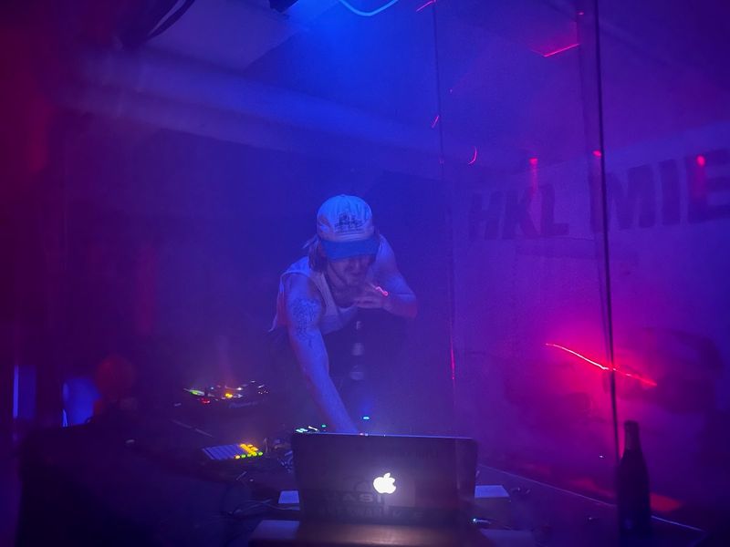 96 back is on top of a table in a room lit with fluorescent blue and red colors. He holds a cigarette in his left hand reaches to the laptop that runs the audio for the performance. The fog machine is working hard, and so do the lasers that can be seen on the walls.