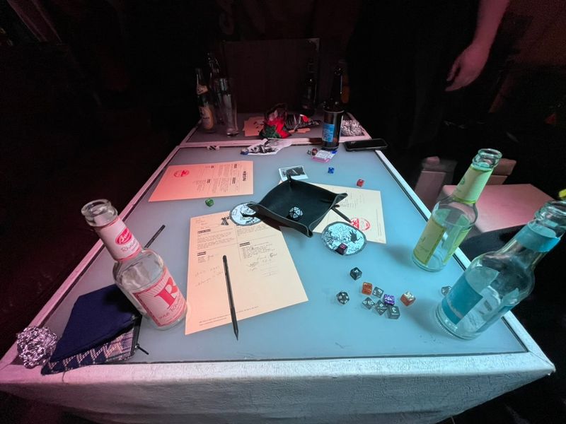 A table at c-base with some artifacts of a TTRPG scattered on top of it: some dice, character sheets and empty bottles of fizzy drinks